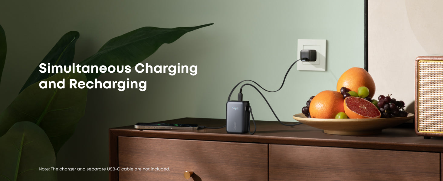 Charging station with laptop and phone on a wooden surface with fruits and a plant.